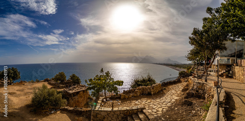 Fototapeta Naklejka Na Ścianę i Meble -  Panoramic view from Atatürk Park's stone-paved observation deck over Antalya Bay with sparkling sea, Konyaaltı beach and hazy mountains under layered clouds. Antalya, Turkey.

