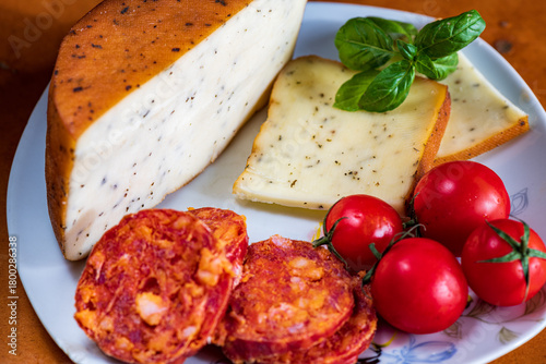 Sliced kulen and semi-hard cheese arranged neatly on a plate, garnished with fresh basil leaves. The vibrant colors of the red cured sausage, creamy-yellow cheese, and green basil create a visually ap