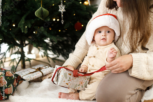 Happy family opening stylish christmas gifts under decorated festive tree, close up. Merry Christmas and Happy Holidays ! Mother and baby son in santa hat unpacking together xmas presents