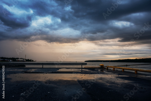 Dramatic coastal landscape with dark storm clouds, sunset light and wet asphalt reflecting the sky. Moody seascape with harbor elements, calm water and a distant shoreline under heavy weather.