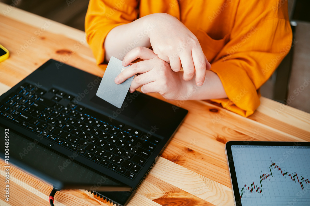 Obraz premium woman inorange shirt is seated at a wooden desk holds credit card making online purchases on laptop, tablet with graph is visible nearby
