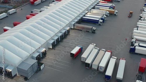 Aerial view of a modern logistics warehouse with multiple semi-trailers trucks parked at ramps for loading and unloading goods