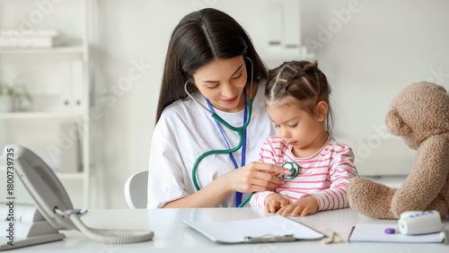 Female pediatrician with stethoscope listening to cute little girl at table i...