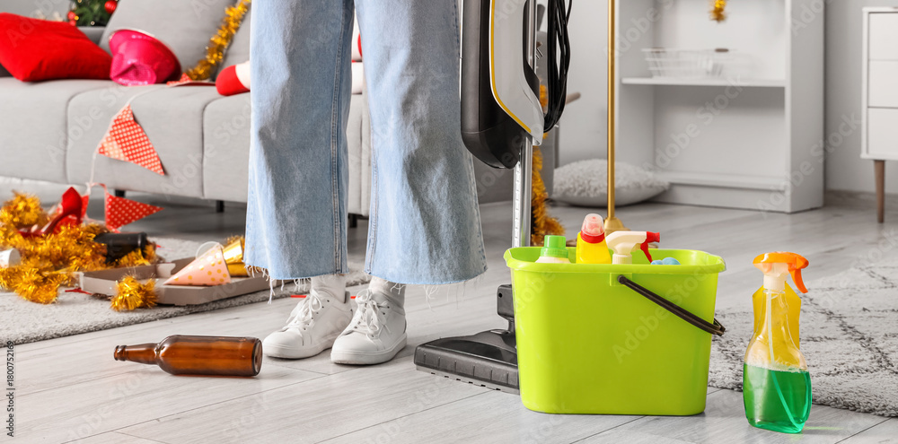 Fototapeta premium Female janitor with cleaning supplies in messy living room after New Year party