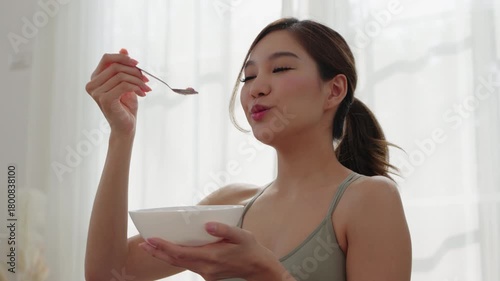 Asian woman in sportswear sitting on yoga mat, enjoying fruit yogurt after workout with morning light and healthy lifestyle mood.