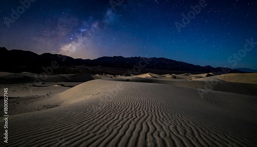 Fototapeta Naklejka Na Ścianę i Meble -  A nocturnal view of desert sand dunes under a starry sky. The Milky Way galaxy glows above dark mountain silhouettes