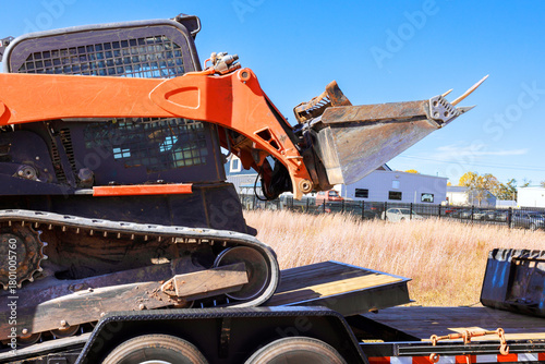 Skid steer machinery loader is positioned on flatbed trailer under its equipment.