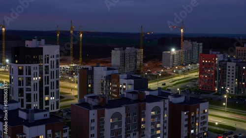 Top view of the construction site. Night timelapse of working cranes, construction of an apartment building. Glare from passing cars, lights turned on in the windows of houses.