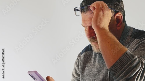 Elderly man struggles to read a message on his smartphone while wearing glasses indoors