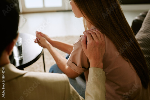 Caucasian young adult woman sitting with hands clasped while middle aged man placing hand on her shoulder providing support during psychological counseling session