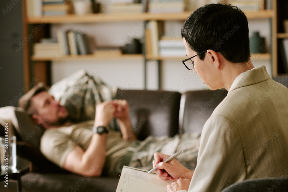 Fototapeta premium Middle aged Caucasian man lying on couch talking, while middle aged woman psychologist sitting in foreground writing notes during therapy session in office setting