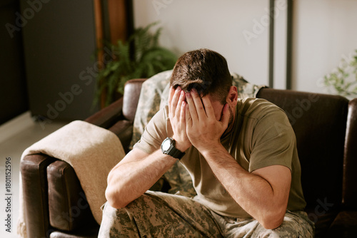 Foto Caucasian young adult man in military uniform sitting on couch covering face wit