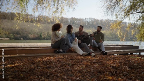 A group of friends shares laughter and stories on a lovely autumn day by the river, surrounded by colorful leaves and warm sunlight.
