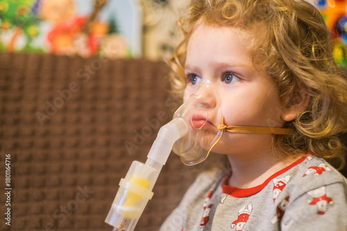 Little girl with curly hair uses nebulizer mask. Child with respiratory illness having inhalation therapy at home for asthma or bronchitis.