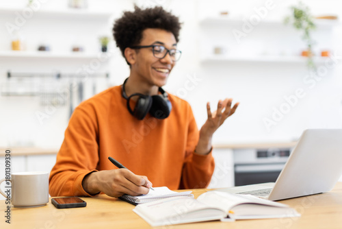 A young man with glasses smiles while working on his laptop, taking notes in a notebook, and wearing headphones.