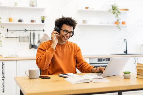 A young man with glasses and headphones smiles while working on his laptop at a wooden table in his kitchen.