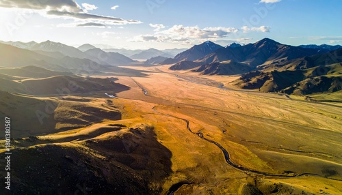 An aerial view captures a vast, sun-drenched valley with rolling hills, a meandering river, and majestic mountains under a partly cloudy sky.