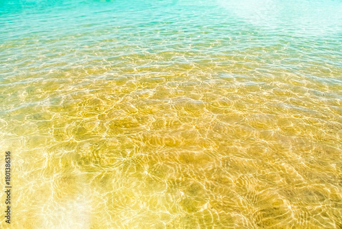 A background of clear water over a sandy bottom. Shallow water with yellow sand. Gentle ripples on the surface. Glittering reflections on the sea.