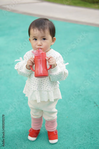 A toddler in a white dotted outfit holds a red sippy cup on a green playground surface