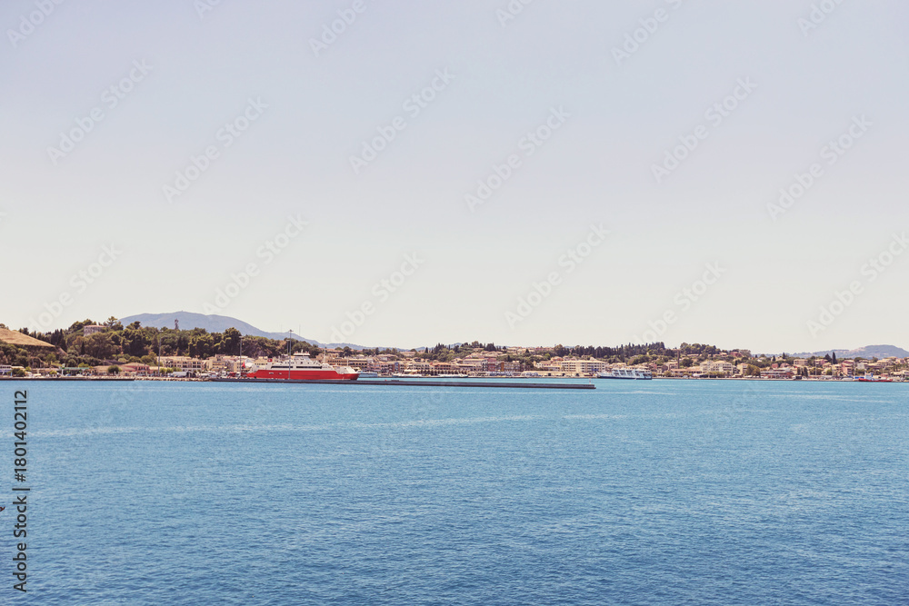 Fototapeta premium Corfu sunlit shoreline and deep blue sea frame docked ships and distant buildings, Greece