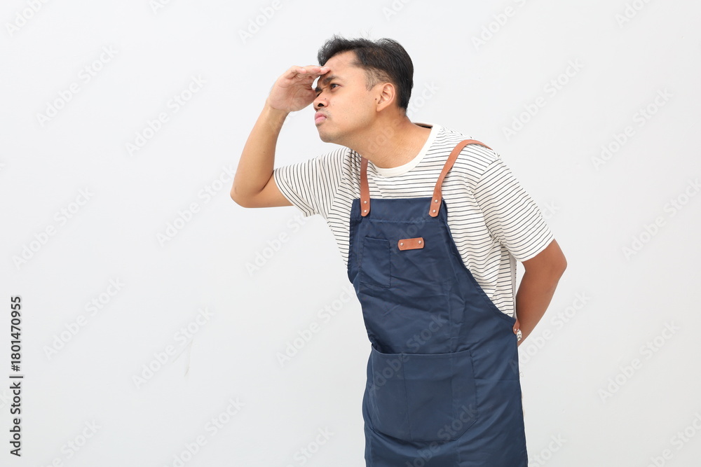 Fototapeta premium Portrait of a curious Asian man in an apron, holding his hands over his eyes and peering into the distance, looking far away, expecting and searching for someone, isolated on a white background.