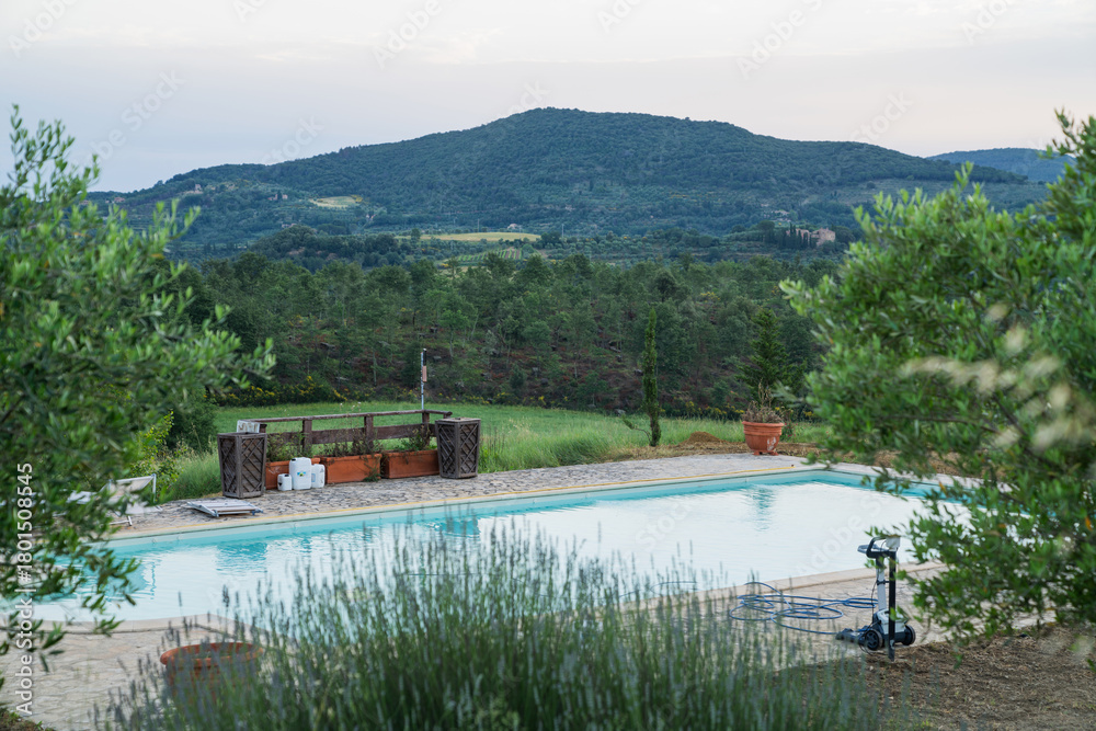Fototapeta premium Swimming pool with clear water on a stone terrace overlooking olive trees, wildflowers, and forested hills during sunset in the countryside of Tuscany, Italy, with soft light over the landscape.