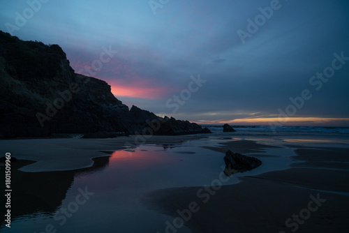 A serene coastal landscape at twilight with rugged cliffs silhouetted against a sky painted in hues of pink and blue. The calm waters gently reflect the colors of the setting sun.