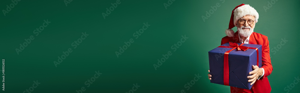 Naklejka premium Handsome senior man in a festive red suit holds a gift, celebrating winter cheer and joy