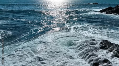 Dynamic Sea Waves Swirling Near Rocky Shoreline with Bright Sun Glare on Deep Blue Water