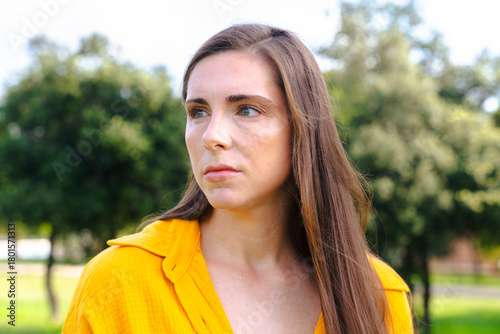 Portrait of serious young woman wearing yellow shirt, looking away thoughtfully in green park.Calm environment and emotional expression concept representing reflection and introspection.
