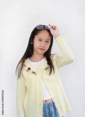 A cute young Asian girl in a yellow cardigan and jeans against white background is posing while adjusting her sunglasses on her head.
