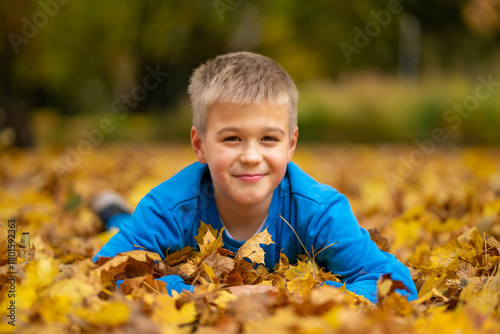 A cute smiling boy in a blue jacket lies on the ground among fallen autumn leaves