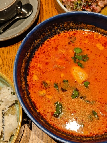 Thai tom yum soup with rich red broth herbs, and coconut base served in a blue bowl. Perfect stock photo for restaurant menus, food packaging, advertising campaigns.