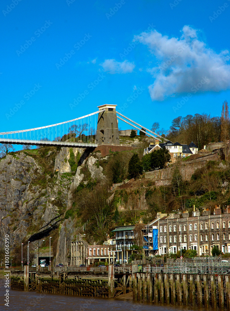 Fototapeta premium View of the Clifton Suspension Bridge in Bristol, England, with buildings and a river in the foreground.