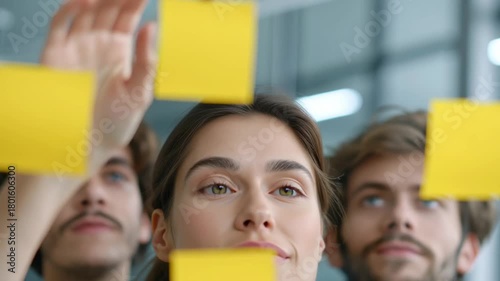 A woman raises her hands toward a glass surface while placing yellow sticky notes, slightly adjusting their position as colleagues observe behind her, creating a focused and creative planning moment
