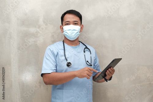 Southeast Asian male healthcare worker in scrubs and face mask using tablet, isolated on light wall, ideal for medical ads or safety campaigns.
