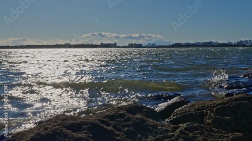 A bright, shimmering reflection of sunlight on the dark, choppy bay water, with rugged rocks framing the bottom foreground.