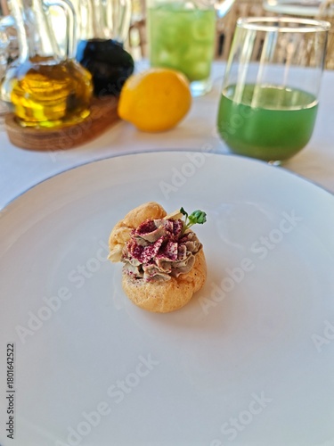 Elegant restaurant appetizer — choux pastry filled with liver pâté, garnished with herbs and beetroot powder. Fine dining amuse-bouche served on a white plate with drinks and olive oil in the backgrou