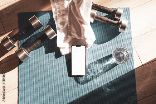 Smartphone mockup on towel with dumbbells, glass of water, and yoga mat in sunlight. Home fitness setup for online workouts, self-care, remote training, and healthy lifestyle.