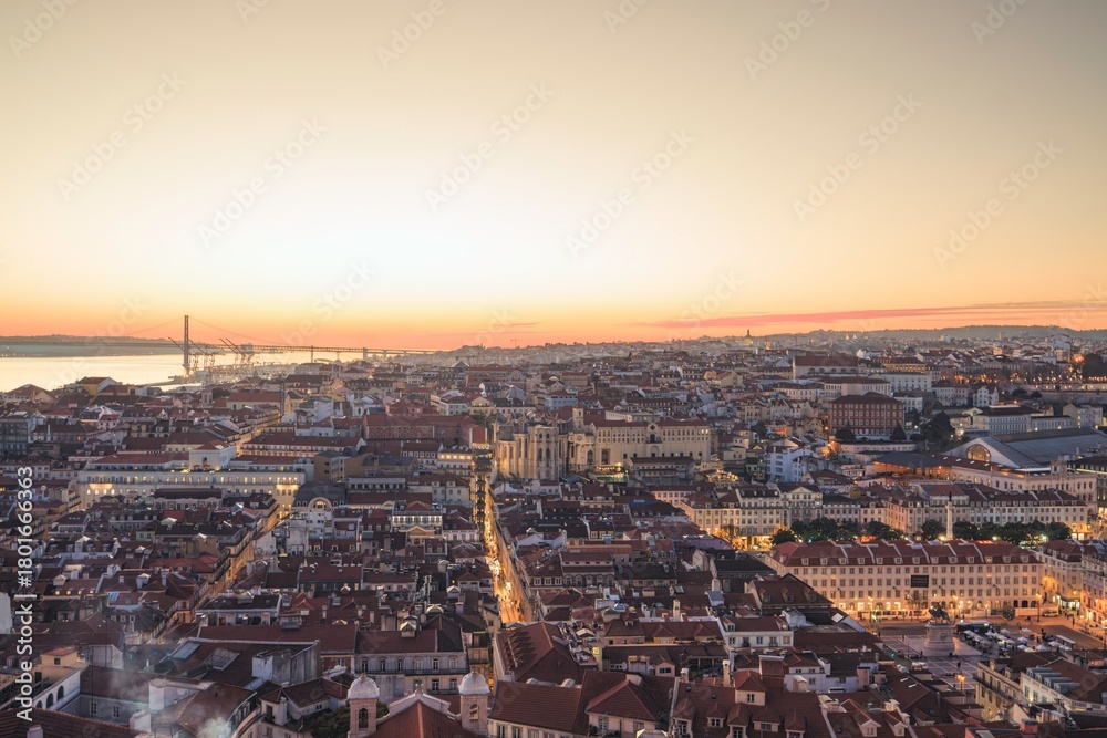 Fototapeta premium As the sun sets, Lisbon's rooftops glow in warm hues. The river reflects the fading light, while people below enjoy the evening atmosphere, bustling with life