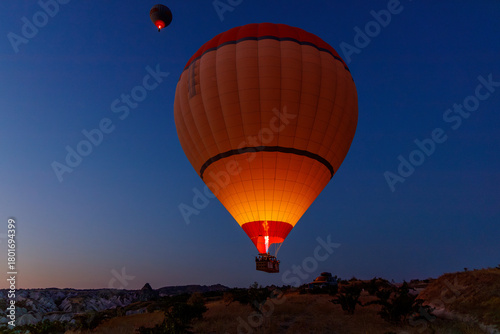 Two hot air balloons glow against a deep blue twilight sky. One large, orange-red balloon dominates the frame, with a smaller, distant one above. Cappadocia, Turkey