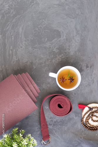 Flat lay featuring a rolled yoga mat, a yoga strap, a wooden mala beads, and a cup of herbal tea with star anise. Holistic wellness and mindfulness of a yoga or Ayurveda practice.
