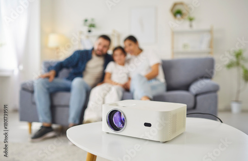 Family watching movie with projector at home. Focus on modern device on white coffee table, with parents and child girl relaxing on sofa in living room. Cozy home cinema, family togetherness.