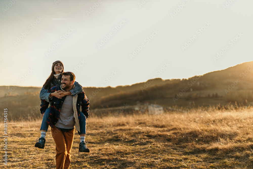 Fototapeta premium Smiling couple piggybacking in nature during sunset