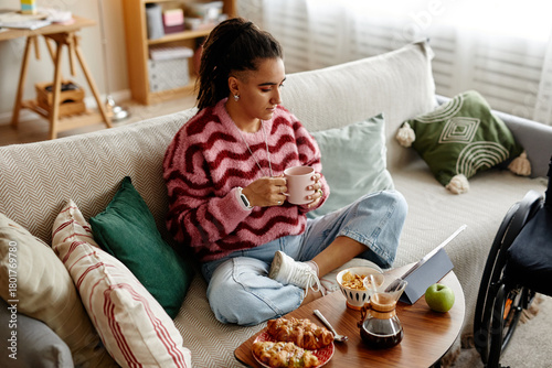 Slika na platnu Young adult woman sitting cross legged on sofa holding mug and looking at laptop