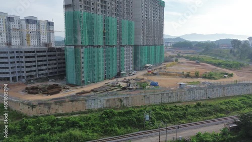 Drone fly-up revealing a massive high-rise apartment construction site wrapped in green safety netting. Active work area below, next to railway tracks and overgrown slopes.