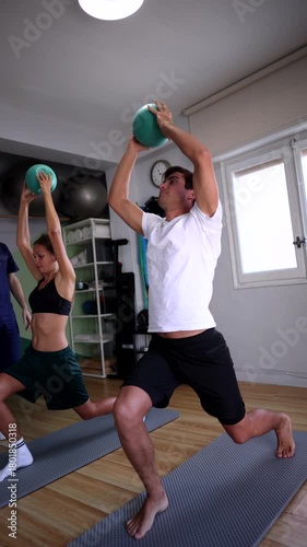 Fit young couple doing synchronized lunges while holding small exercise balls in a modern gym, enhancing their strength, balance, and coordination during a functional training session