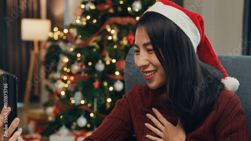 Happy Asia girl wear red santa hat new normal celebrate christmas party at home with smartphone online virtual event video call conference with friend an family on wing chair in living room at house.