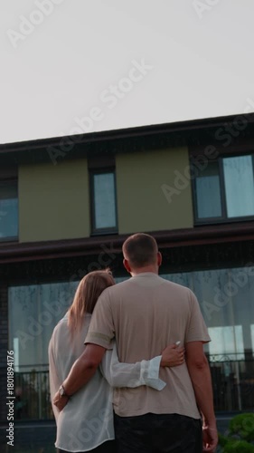 Young couple embraces in the rain, gazing at their contemporary house, representing their joint aspirations, investment in real estate, and promising future in their new home