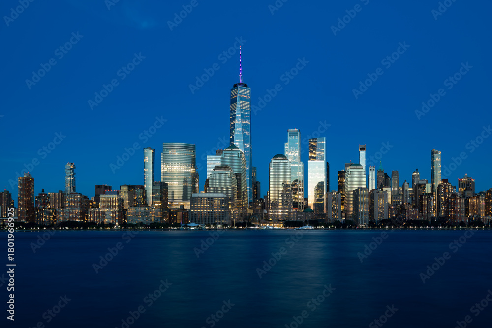 Fototapeta premium A stunning nighttime view of the downtown Manhattan skyline, seen across the Hudson River from Jersey City, with the towers of the Financial District glowing over the dark, tranquil water.